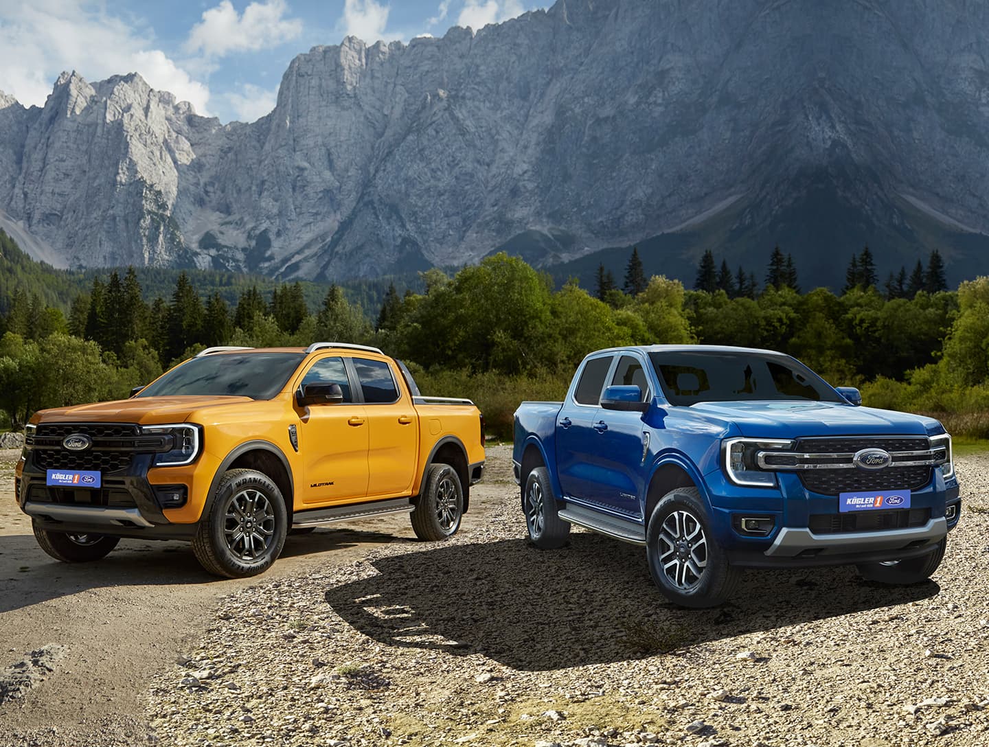 Zwei Ford Pickups, einer gelb und einer blau, stehen nebeneinander in einer natürlichen Umgebung mit Bergen im Hintergrund