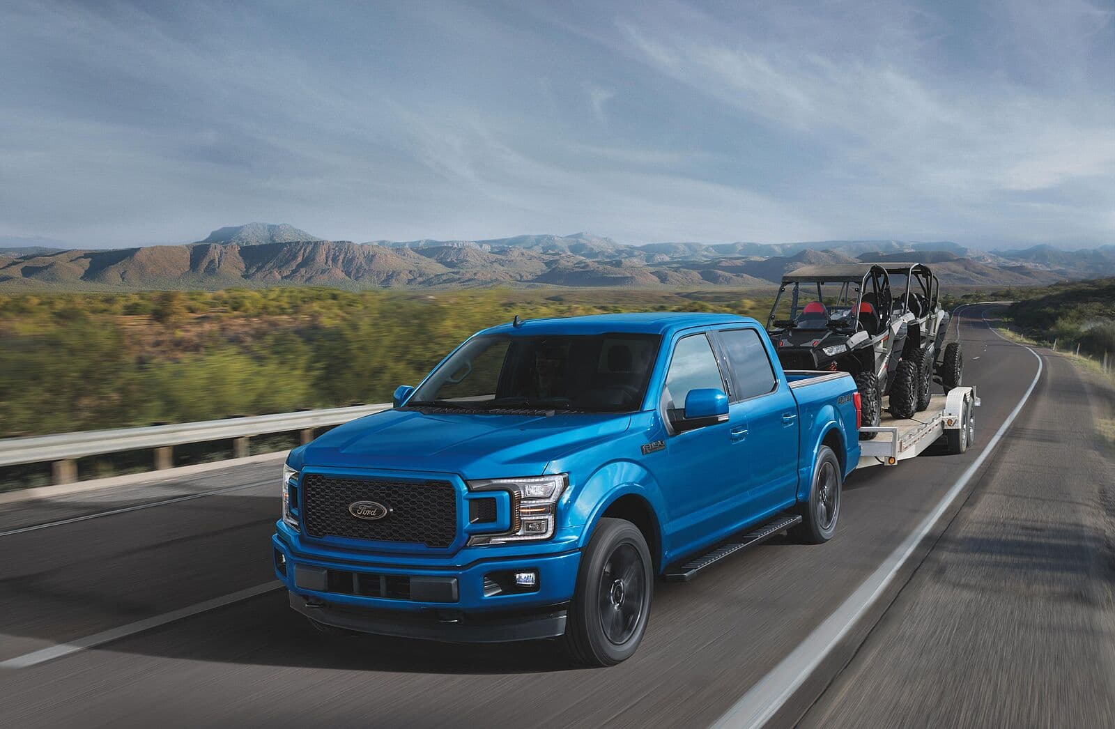 Ford F-150 in Blau auf einer Straße, Anhänger mit zwei Offroad-Fahrzeugen im Hintergrund, Berge im Hintergrund