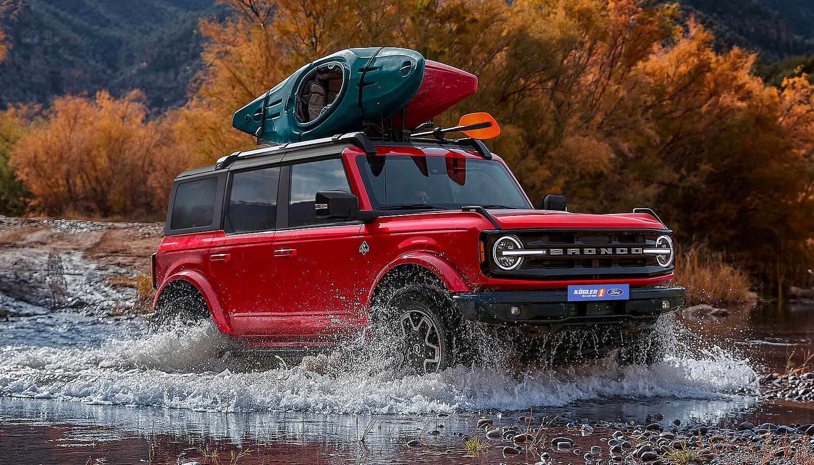 Ford Bronco in roter Farbe fährt durch Wasser mit einem Kajak auf dem Dach, umgeben von herbstlichen Bäumen